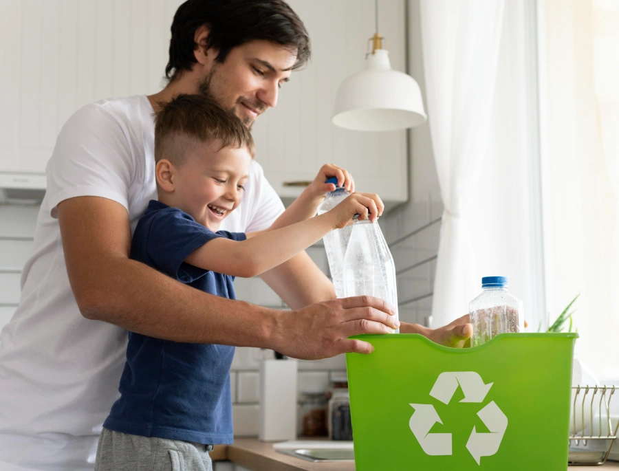 Un padre y su hijo sonríen mientras juntos colocan botellas de plástico en una canasta de reciclaje verde en la cocina