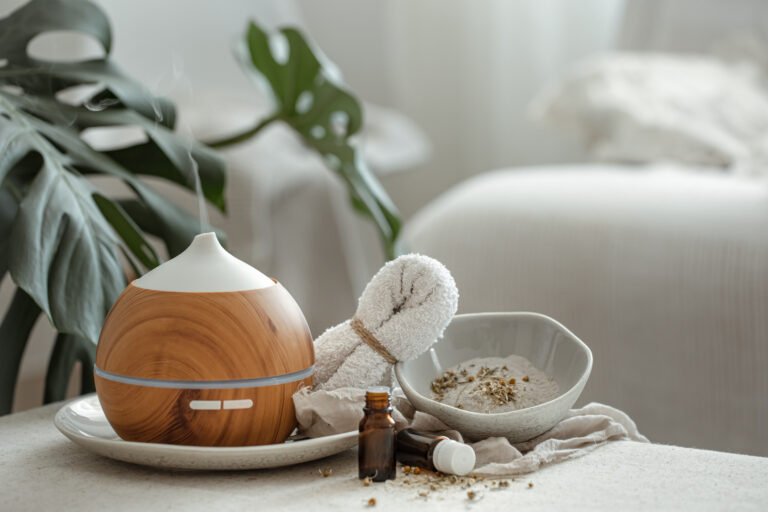 Difusor de aromas con acabado de madera emitiendo vapor junto a frascos de aceites esenciales y un cuenco con hierbas.