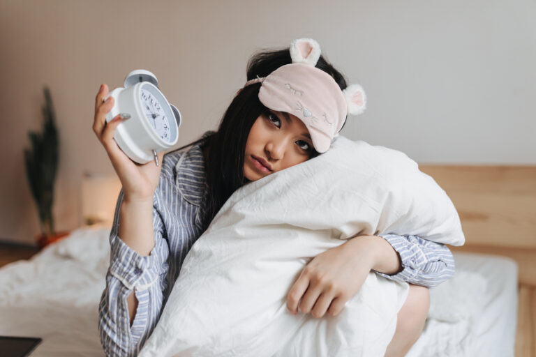 Mujer con aspecto cansado sosteniendo un reloj despertador y una almohada tras una noche de sueño insuficiente.