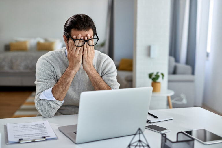 Hombre con gafas cubriendo su rostro con las manos por fatiga frente a una laptop en su casa.