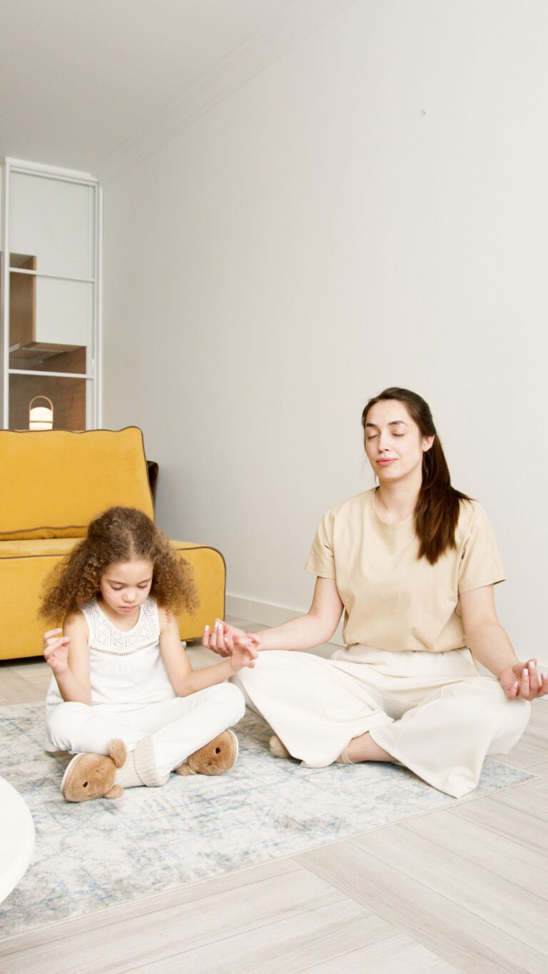 Madre e hija sentadas en el suelo de la sala meditando en postura de loto con los ojos cerrados.