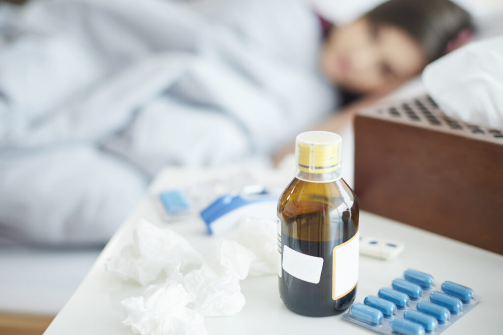 Jarabe para la tos, cápsulas y pañuelos sobre una mesa de noche junto a una persona descansando en cama por síntomas respiratorios.